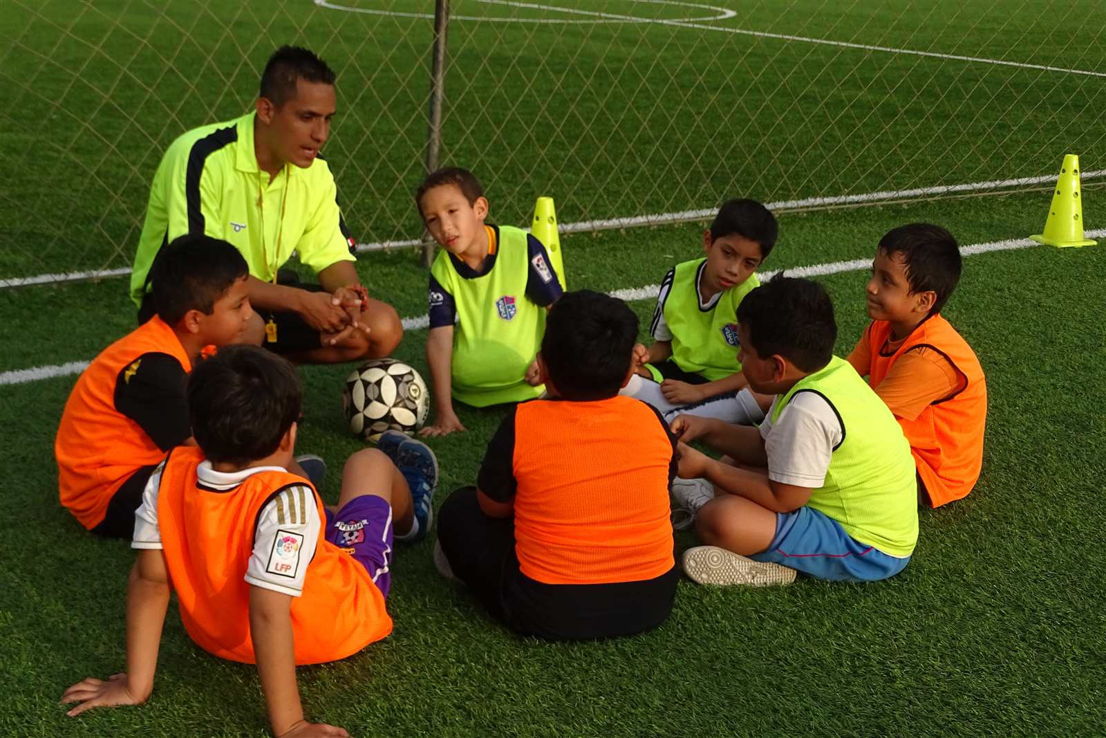 Entrenador con alumnos con petos de colores sentados en círculo en la cancha